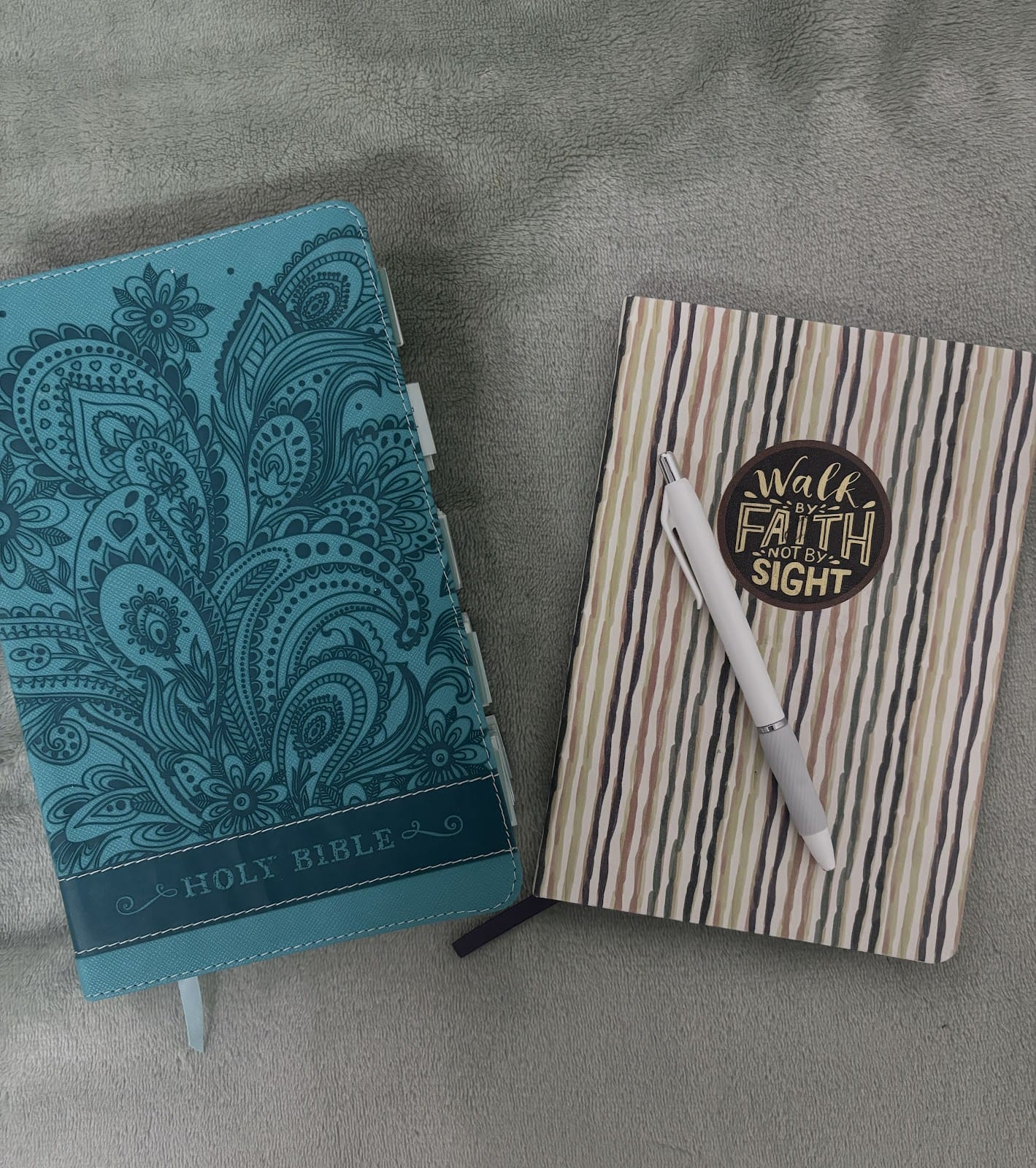  A teal-colored Bible with intricate floral patterns next to a striped journal with a cover that reads, "Walk by faith, not by sight." A pen is placed on top of the journal.