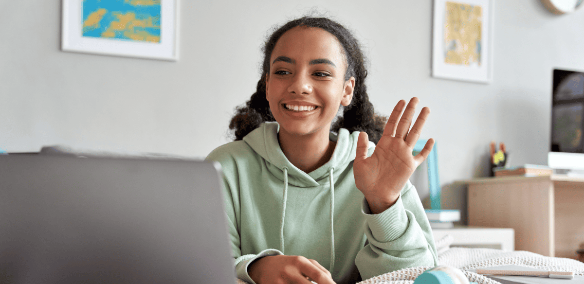 A young woman sits at a desk with her laptop, smiling and waving cheerfully at the camera.