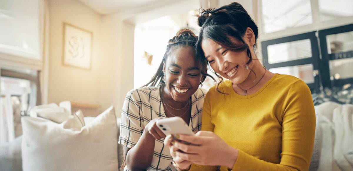 Two women smiling and engaging with their cell phones, sharing a joyful moment together.