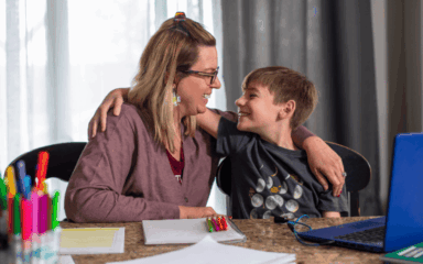 A woman and a boy are seated at a table, engaged with a laptop in front of them.