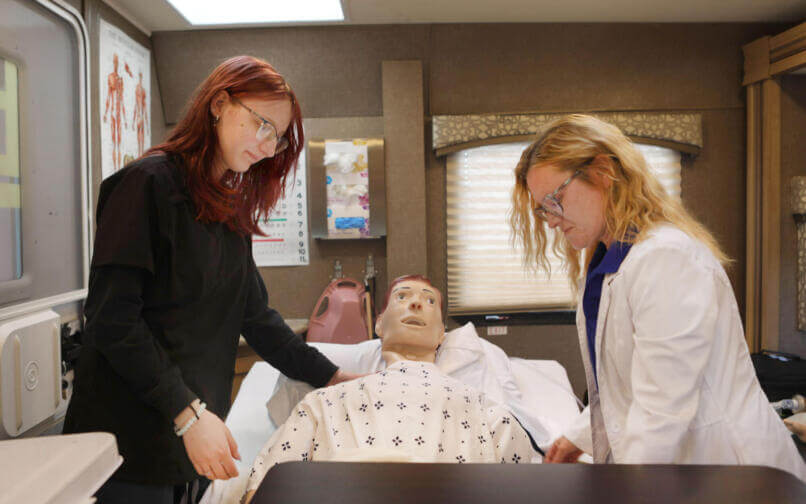 Two healthcare students practice patient care on a medical training mannequin in a simulation lab.