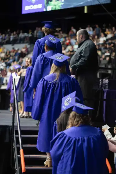 Graduates in blue robes ascending a stage at a ceremony, wearing decorated caps. A crowd watches in the background, creating a celebratory atmosphere.