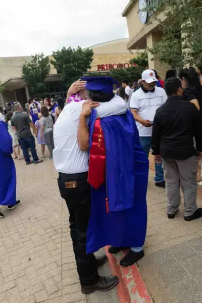 A graduate in a blue gown and cap embraces an older adult in white outside a venue. The scene is joyful, with a crowd celebrating in the background.