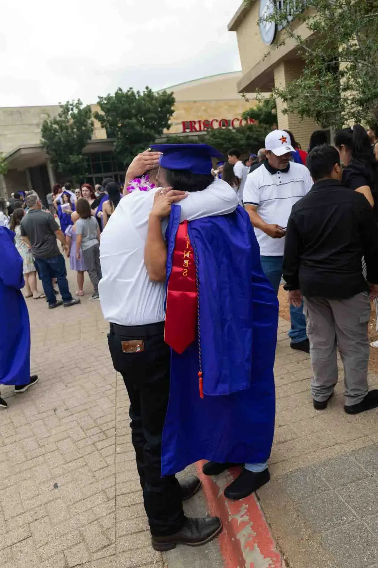 A graduate in a blue gown and cap embraces an older adult in white outside a venue. The scene is joyful, with a crowd celebrating in the background.