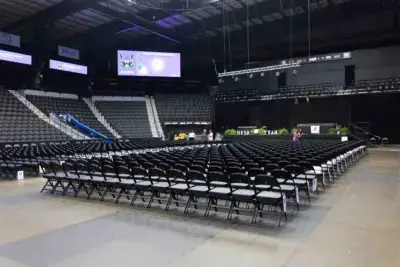 An empty indoor arena with rows of black chairs facing a stage. Large screens display logos, and green plants decorate the stage, creating an anticipatory atmosphere.