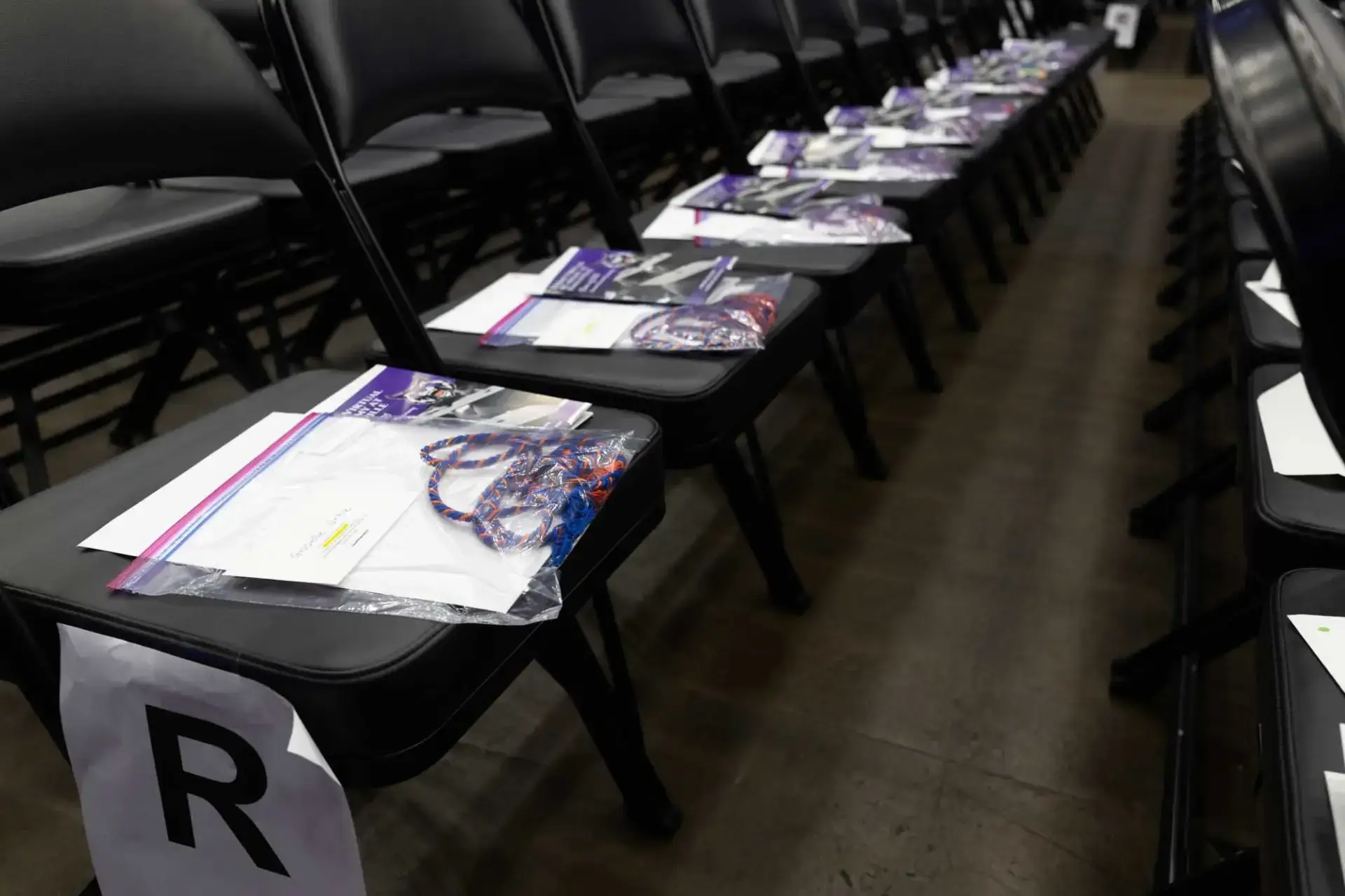 Rows of black chairs with packets on each seat, including a booklet and a colorful cord. A paper labeled "R" marks a row. The setting is calm and organized.