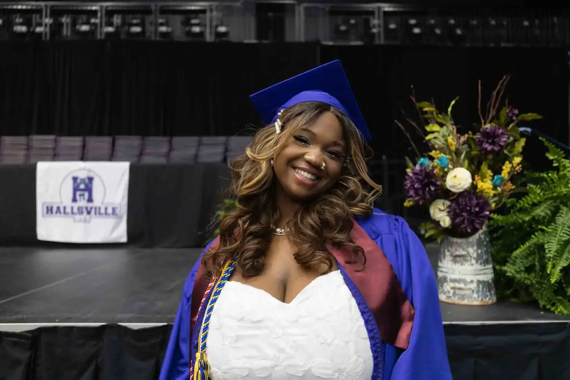 Smiling graduate in blue cap and gown stands on stage with Hallsville banner in the background. A bouquet of colorful flowers is nearby, conveying joy.