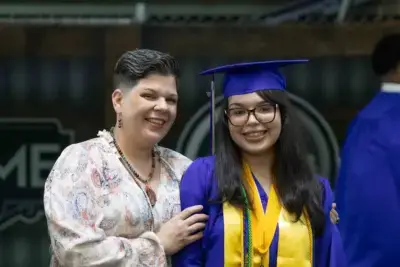 A smiling graduate in a blue cap and gown stands beside a proud woman with short hair. The scene conveys joy and accomplishment.