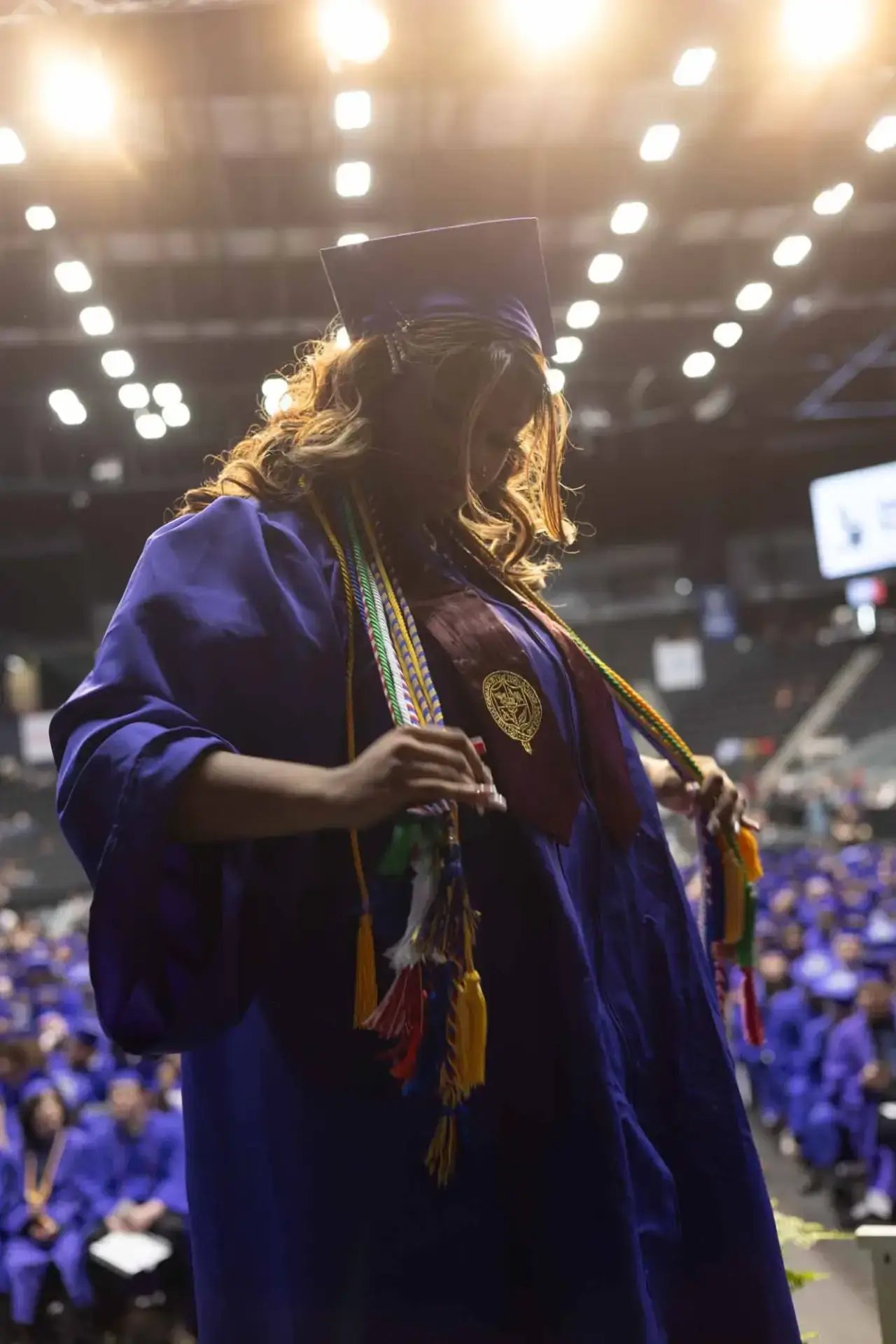 A graduate in a blue cap and gown stands holding colorful honor cords in a brightly lit auditorium filled with peers, conveying pride and accomplishment.