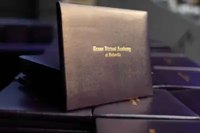 Stack of dark diploma covers with gold text reading "Texas Virtual Academy at Hallsville," symbolizing graduation and achievement.