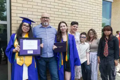 Two graduates in blue caps and gowns hold diplomas, smiling proudly. They're surrounded by supportive family members against a brick wall backdrop.
